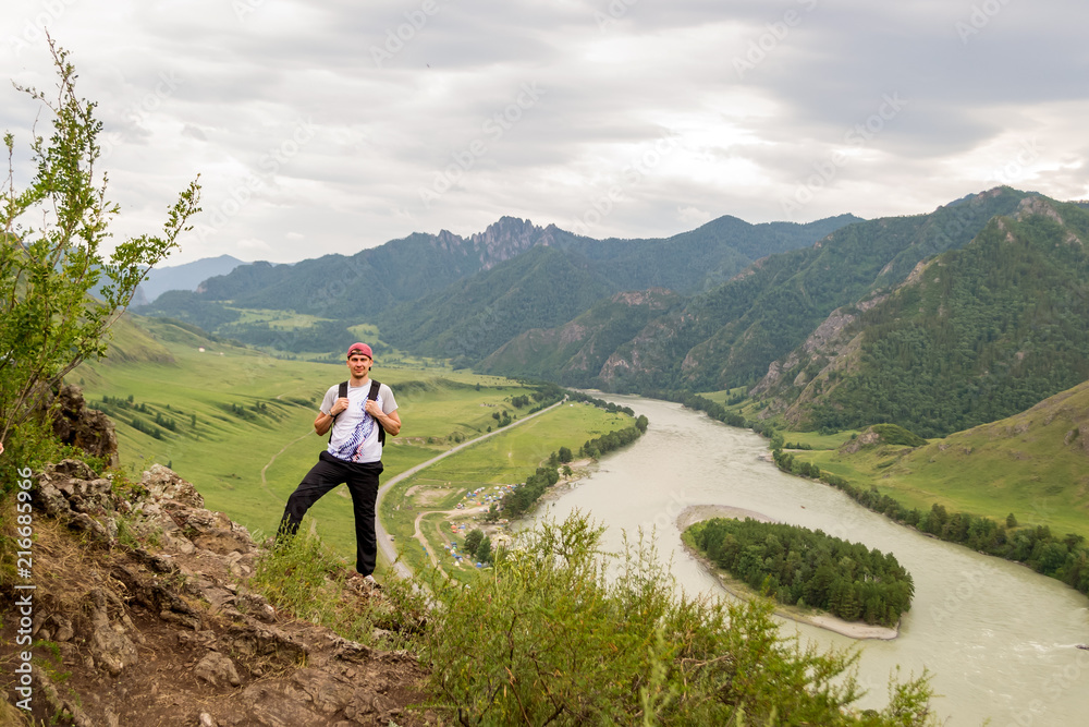 Obraz premium A young guy in a cap with a backpack and black sports pants is standing on the edge of a cliff in the mountains of the Altai, from where he opens a view of the river Katun and the island