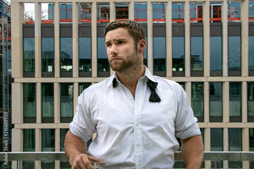 Handsome hunky man in tuxedo, unbuttoned shirt and tie stands on hotel balcony, drinking a beverage photo