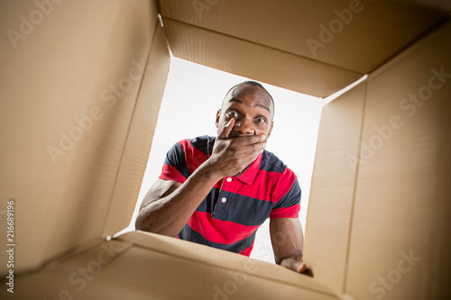 The surprised afrocan man unpacking, opening carton box and looking inside. The package, delivery, surprise, gift lifestyle concept. Human emotions and facial expressions concepts © master1305