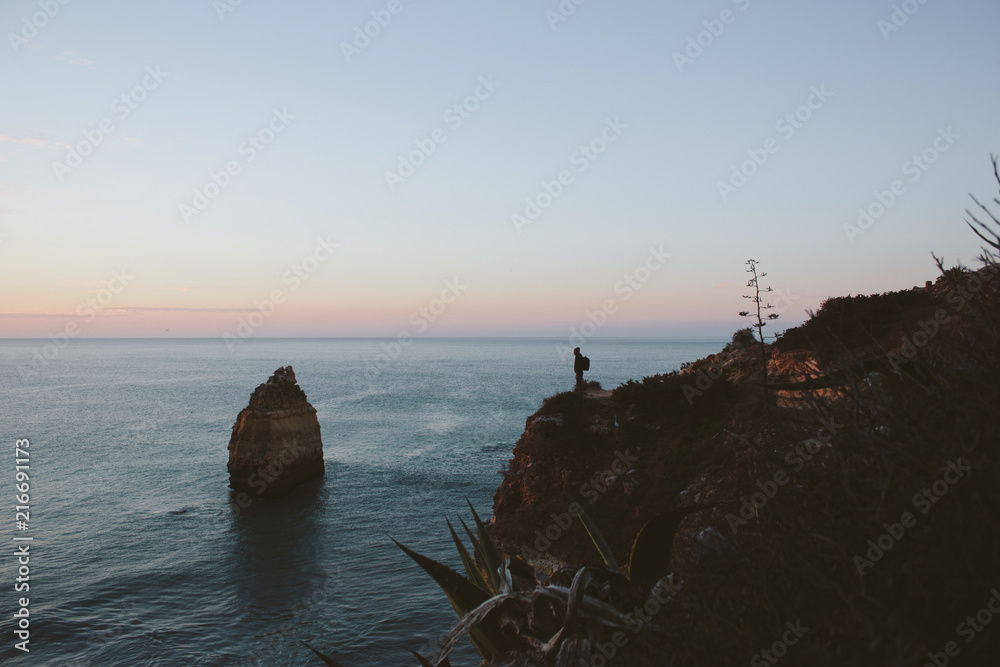 Mid distance view of silhouette man standing on cliff by sea against sky during sunset Mid distance view of silhouette man standing on cliff by sea against sky during sunset