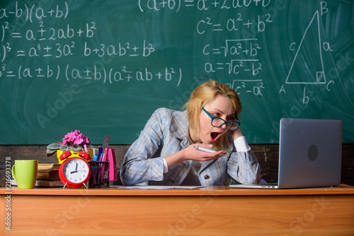 Work far beyond actual school day. Teacher busy with paperwork and research. Teacher tired face keep working after classes. Still working. Teacher woman sit table classroom chalkboard background