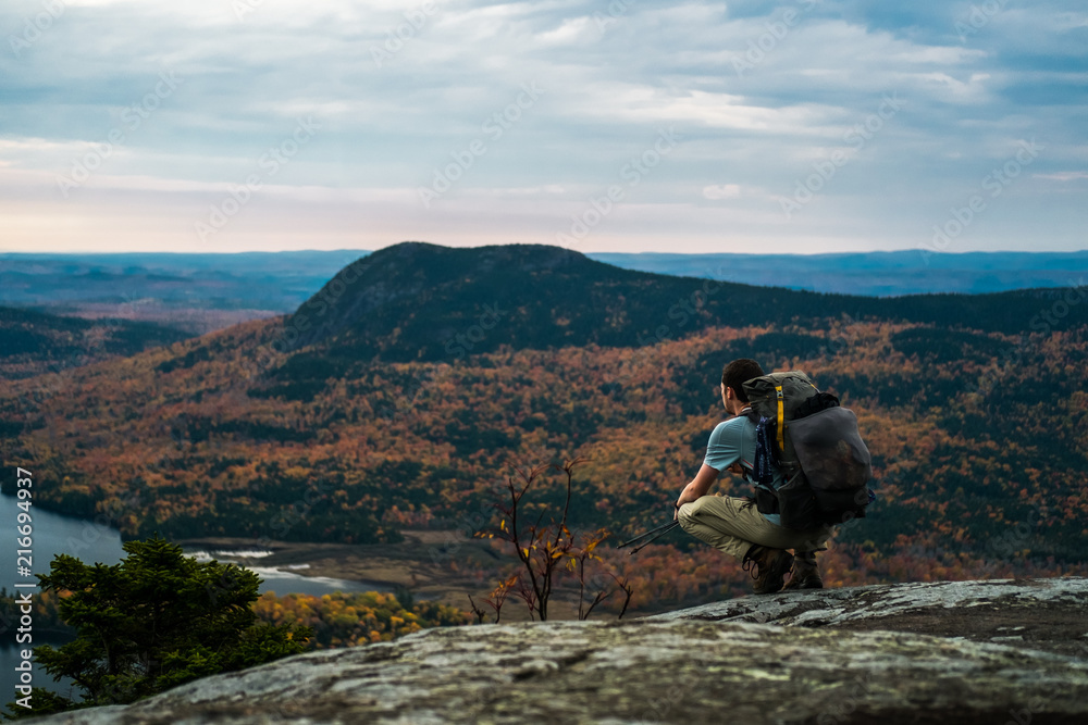 A man with a backpack kneels on top of a mountain in Maine in the fall with autumn foliage and a lake below while backpacking and thru hiking the Appalachian Trail