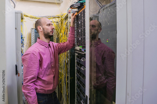 The system administrator works in the server room. The network engineer switches the wires in the data center. A young man is standing next to the racks with computer equipment