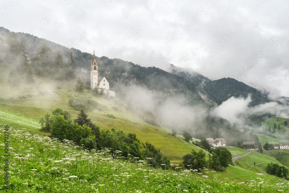 Fototapeta premium Church on a misty hillside with a cloudy valley in the background in the Dolomites mountains in La Valle, South Tyrol, Italy