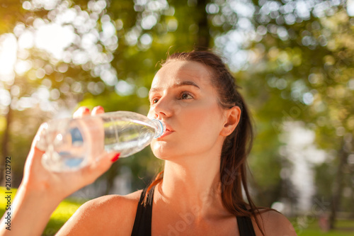 Young sportive woman drinking water after workout in summer park. Healthy way of life