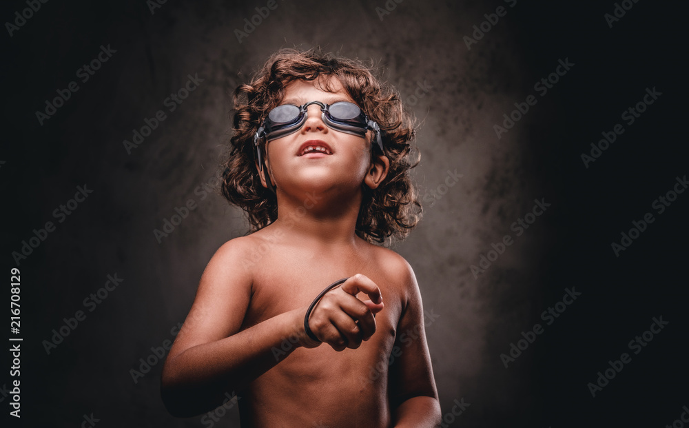 Funny little shirtless boy in swimming goggles in a studio. Isolated on the dark textured background.