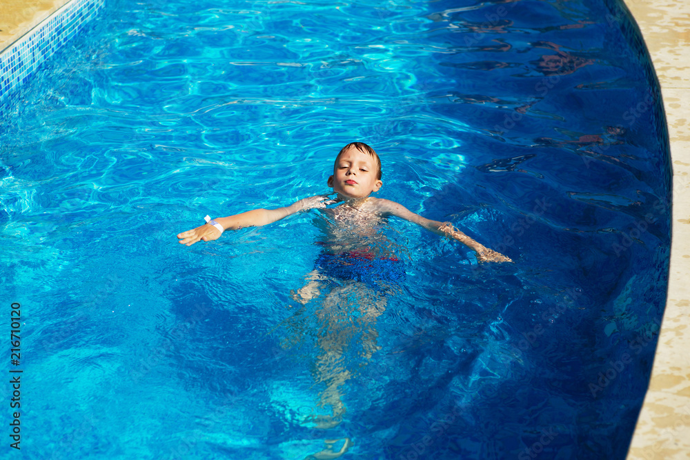 Happy kid playing in blue water of swimming pool on a tropical resort at the sea. Summer vacations concept. Cute boy swimming in pool water