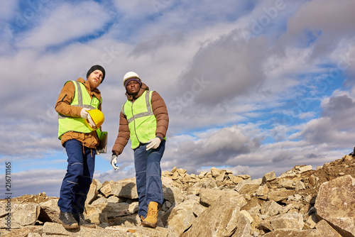 Full length portrait of two industrial workers wearing reflective jackets, one of them African, standing on cliff overlooking mineral mines on worksite outdoors, copy space