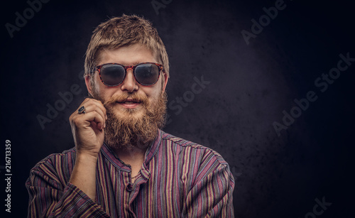 Happy hipster guy wearing sunglasses dressed in an old-fashioned shirt correct his mustache on a dark background.