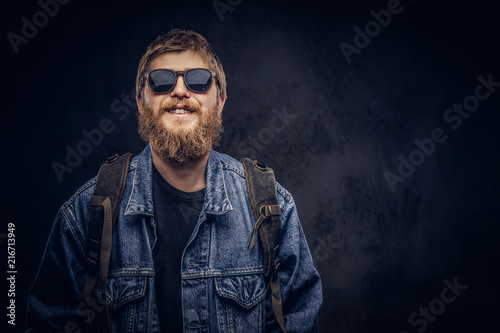 Smiling bearded hipster guy wearing sunglasses and backpack dressed in jeans jacket on a dark background.