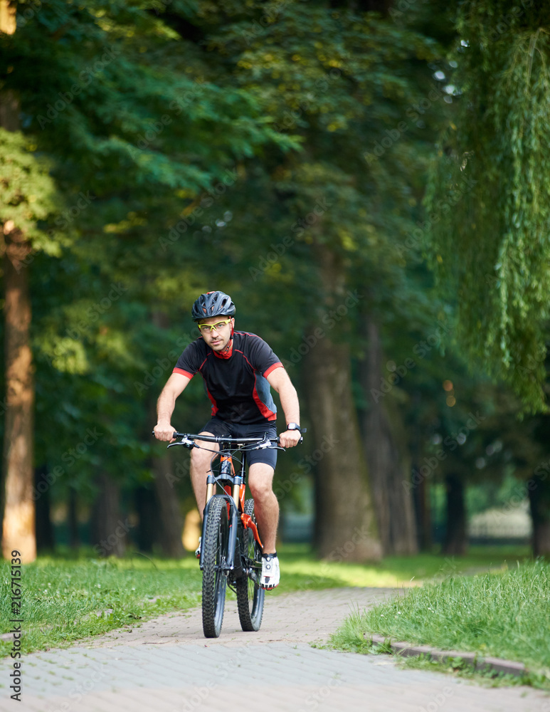 Fototapeta premium Handsome toned guy bicyclist in cycling clothes, looking to the camera, while riding bike down park alley on summer day. Exercising, reaching goal
