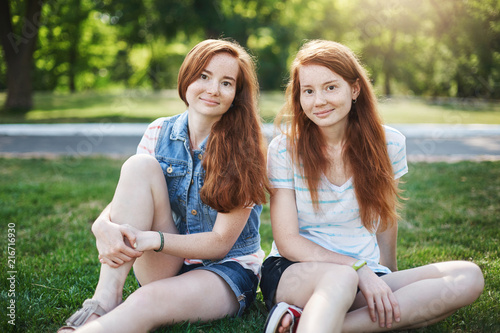 My sister is best friend. Good-looking two female with red hair and freckles, sitting on grass near university campus and chilling with boyfriends, smiling with cute expression while talking casually © Liubov Levytska