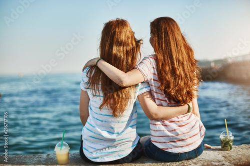 Girs making promises never leave each other, gazing at beautiful sea and hugging, sitting near docks, dreaming about future, talking casually like sisters and enjoying warm sunny evening together photo