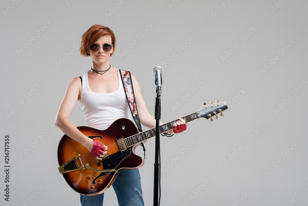 Portrait of a beautiful young rock woman in sunglasses with a guitar. She sings a song in studio microphone Portrait of a beautiful young rock woman in sunglasses with a guitar. She sings a song in studio microphone