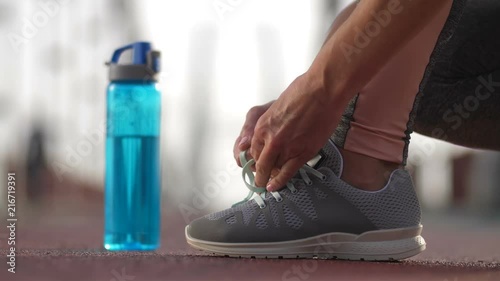 Close-up female runner's feet in sneakers and hands tying shoelaces and taking sport water bottle. Active senior woman running in the morning ties her sports shoes before jogging on bridge. Side view