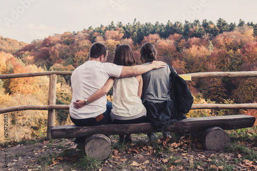 Three people hugged each other sitting on wooden benches and watch the beautiful autumn landscape, concept of polygamy photo