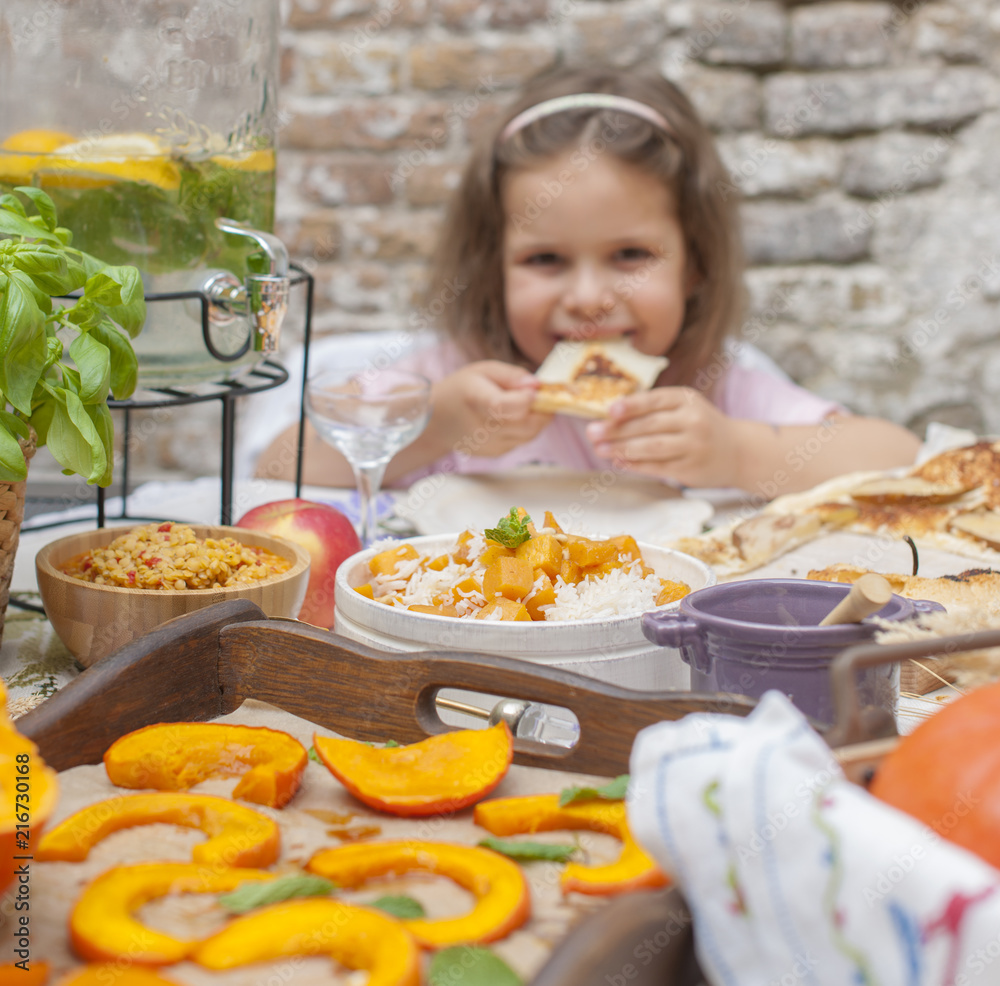 Little girl is eating pie. Family dinner in the courtyard outdoors. Autumn vegetables and fruits on the table. Dining table . A special place for text Little girl is eating pie. Family dinner in the courtyard outdoors. Autumn vegetables and fruits on the table. Dining table . A special place for text