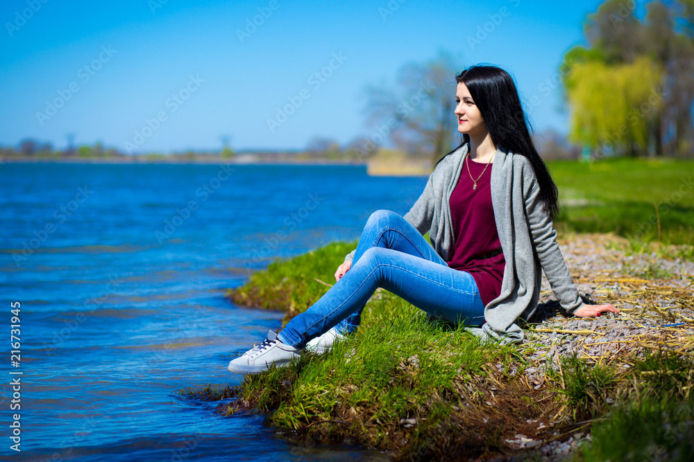 Beautiful young woman in jeans sits on river bank in sunny day