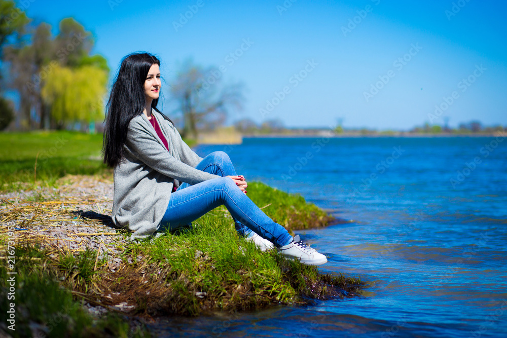 Beautiful young woman in jeans sits on river bank in sunny day