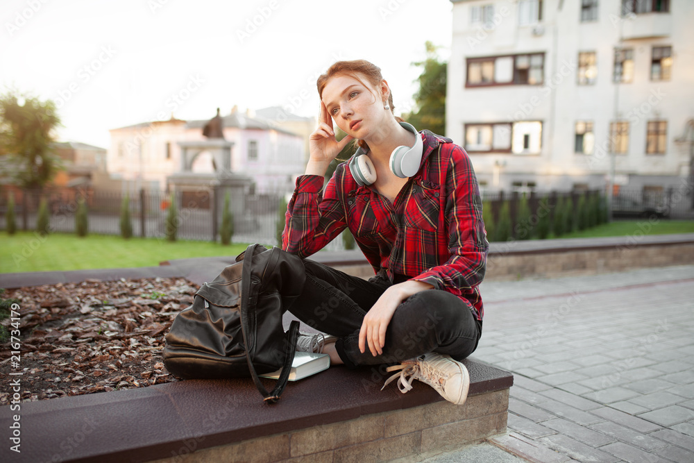 A red-haired girl with freckles is dressed in a red checked shirt is sitting in a lotus pose in the park listening to music on headphones Modern student A red-haired girl with freckles is dressed in a red checked shirt is sitting in a lotus pose in the park listening to music on headphones Modern student