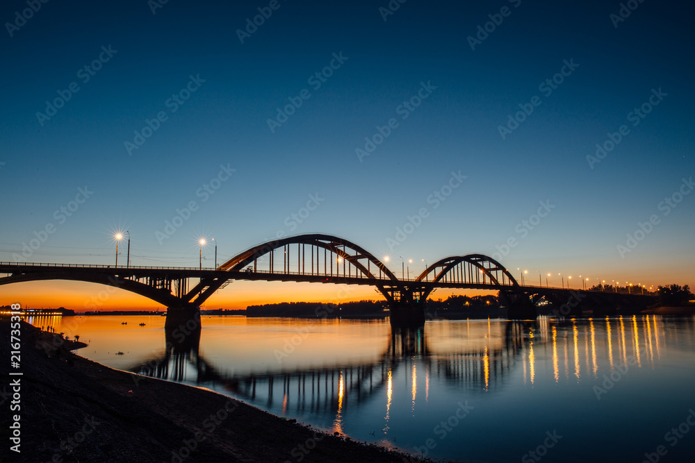 Naklejka premium Volga bridge over Volga river after sunset with reflection in water, Yaroslavl region, Rybinsk city, Russia. Beautiful night landscape