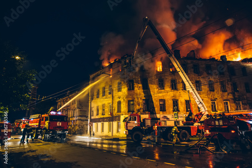 Canvas Print Firefighters with fire hose and fire engines or trucks are fighting fire in residential building at night