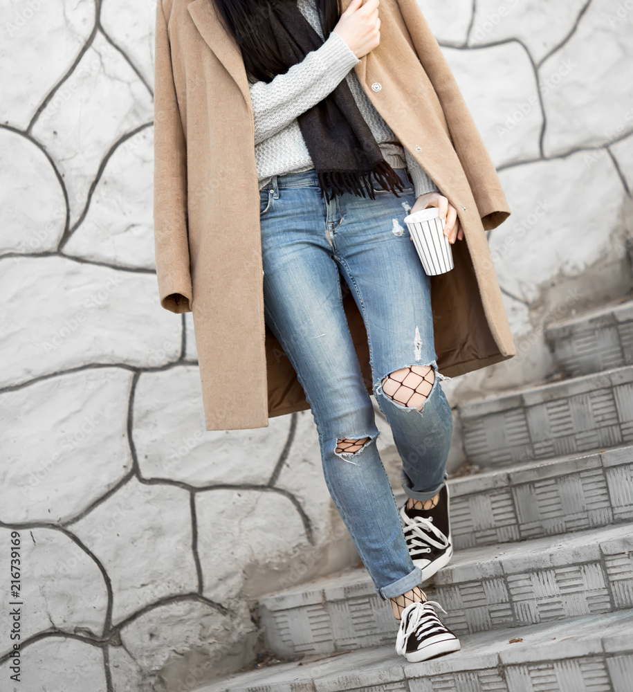 All fashion outfit details. Fashionable woman wearing coat, ripped jeans with sneakers.