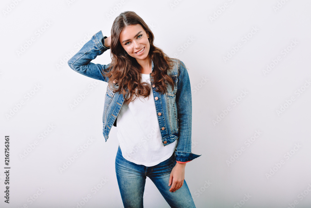 Beautiful young woman with long dark hair dressed jeans suit and white  t-shirt poses with lovely smile on isolated background. Stylish girl with  curly hair stands on the white wall Stock Photo