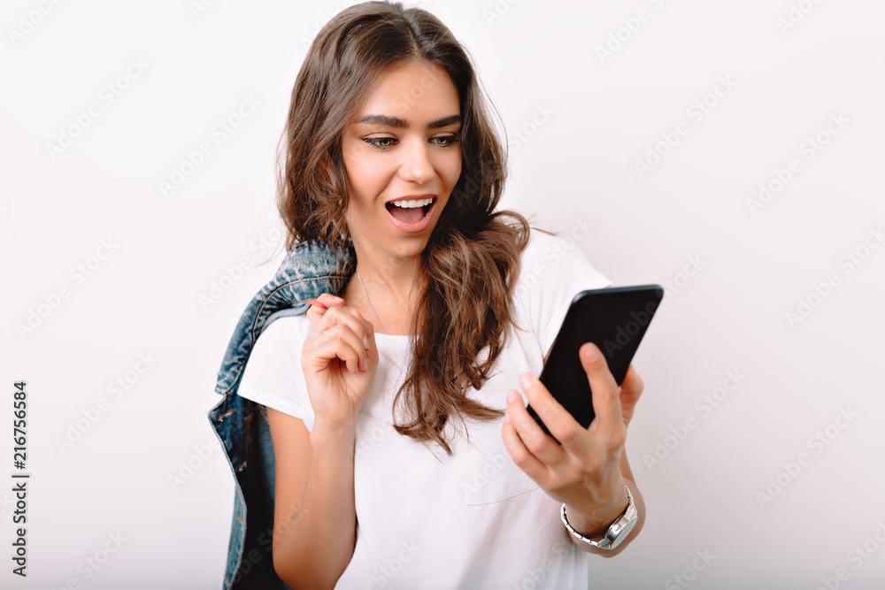 Portrait of pretty woman with long dark hair dressed in white t-shirt looking at smartphone surprised emotions on the white background
