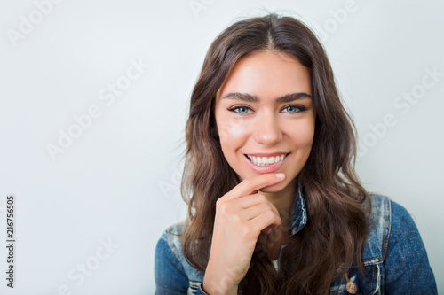 Close-up portrait of sensitive woman with dark hair and big green eyes smiles over grey wall with fantastic smile.