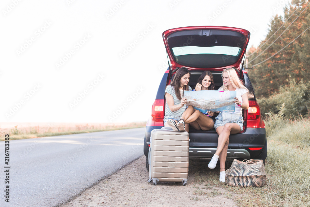 Three young women with suitcases on a trip by car. They sit in the back of the car, they look at the map, resting after a long drive and having fun. Hitchhiking and car trips with friends Three young women with suitcases on a trip by car. They sit in the back of the car, they look at the map, resting after a long drive and having fun. Hitchhiking and car trips with friends