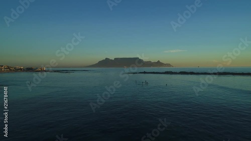 Aerial approach and fly over Bloubergstrand beach with Stand Up Paddle Boarders paddling around on a beautiful evening at sunset with iconic Table Mountain in the background, Cape Town, South Africa. photo