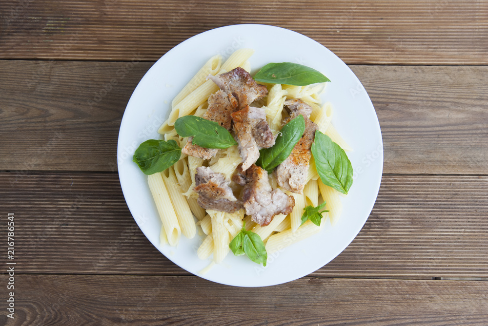 Cooked penne pasta isolated in white round plate, with basil leaves and pork meat, over wooden table, decorated with basil leaves. Top view. Cooked penne pasta isolated in white round plate, with basil leaves and pork meat, over wooden table, decorated with basil leaves. Top view.