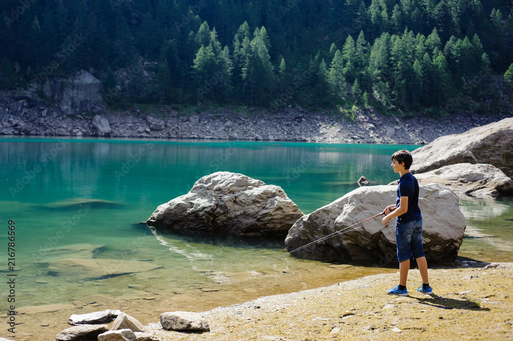 boy fishing in the high mountains in alpine lake, on a summer day, italy piedmont valley ossola