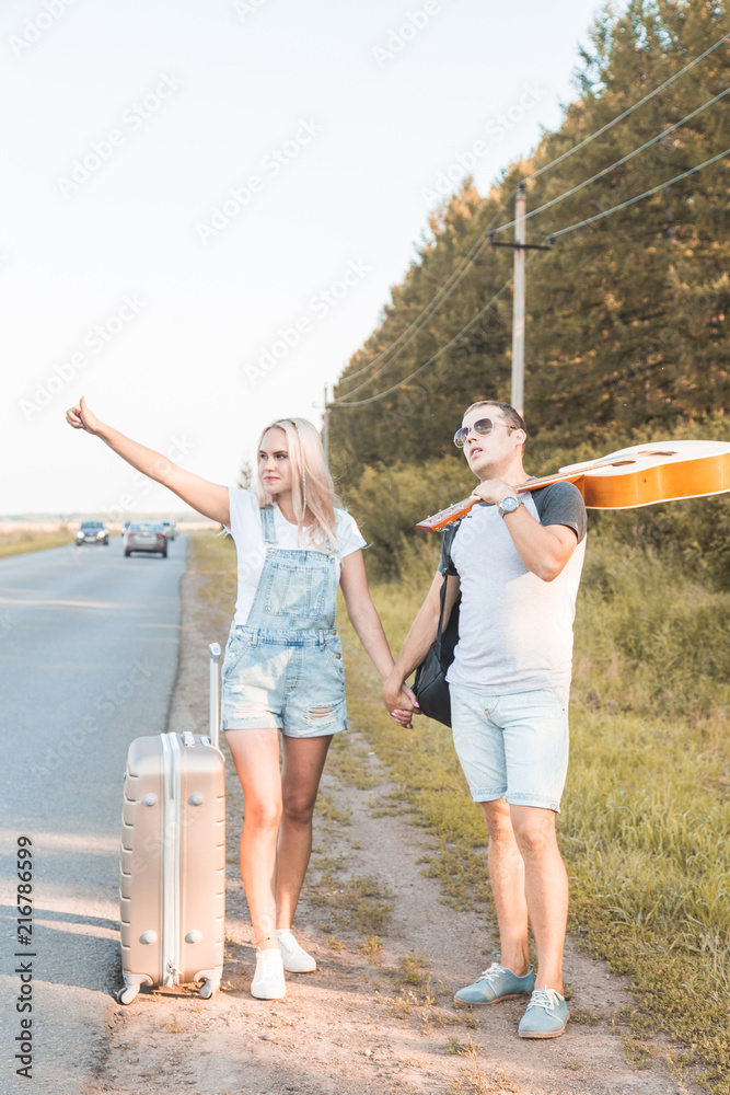 A loving couple walks along the road with a guitar and suitcases and catches a car hitchhiking.