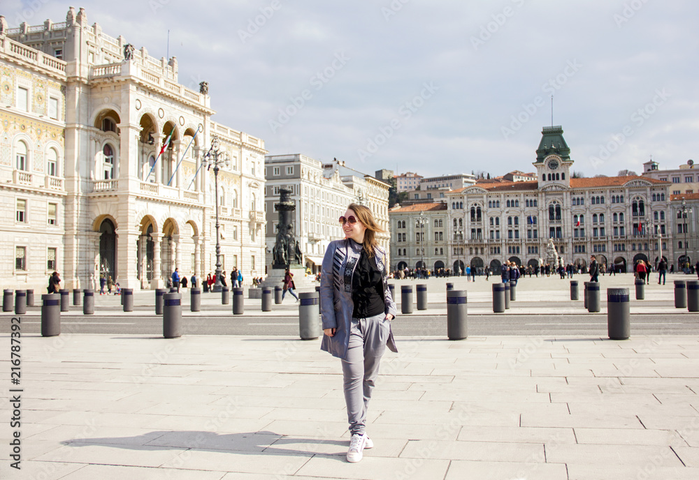 young attractive happy sunny girl in gray pants, cloak and sunglasses is standing fashion with gold backpack and posing on Trieste square background in Italy and smiling