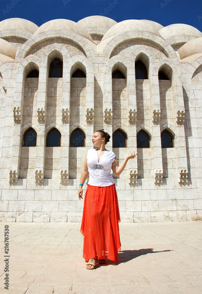 young attractive happy sunny girl in white blouse, long orange skirt and sunglasses is standing like model and posing near white stone church with columns background in Egypt