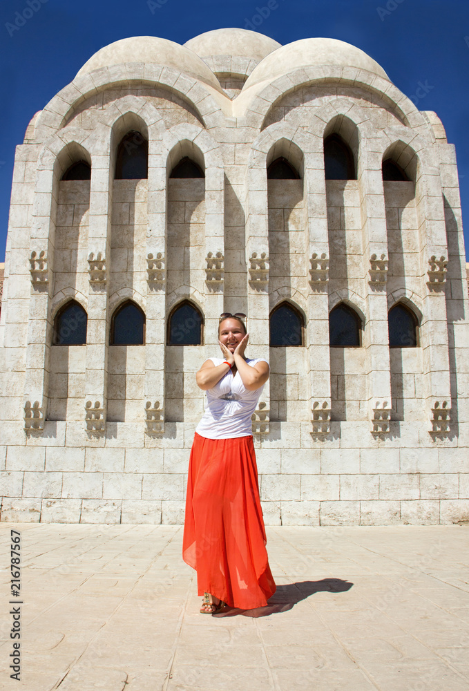 young attractive happy sunny girl in white blouse, long orange skirt and sunglasses is standing like model and posing near white stone church with columns background in Egypt and smiling