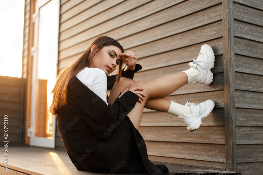 Beautiful young fashionable model woman in black stylish coat with sneakers sitting near a wooden wall at sunset - obrazy, fototapety, plakaty 