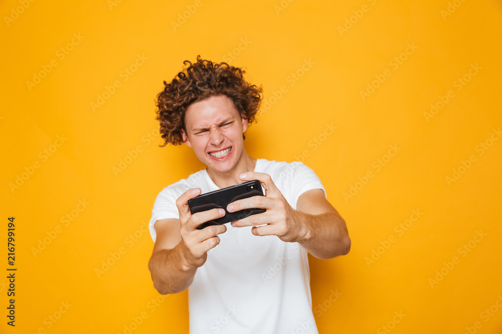 Happy gamer man in casual t-shirt playing online video games on smartphone, isolated over yellow background Happy gamer man in casual t-shirt playing online video games on smartphone, isolated over yellow background