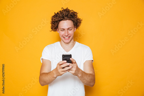 Brunette curly man in casual white t-shirt holding and using mobile phone, isolated over yellow background
