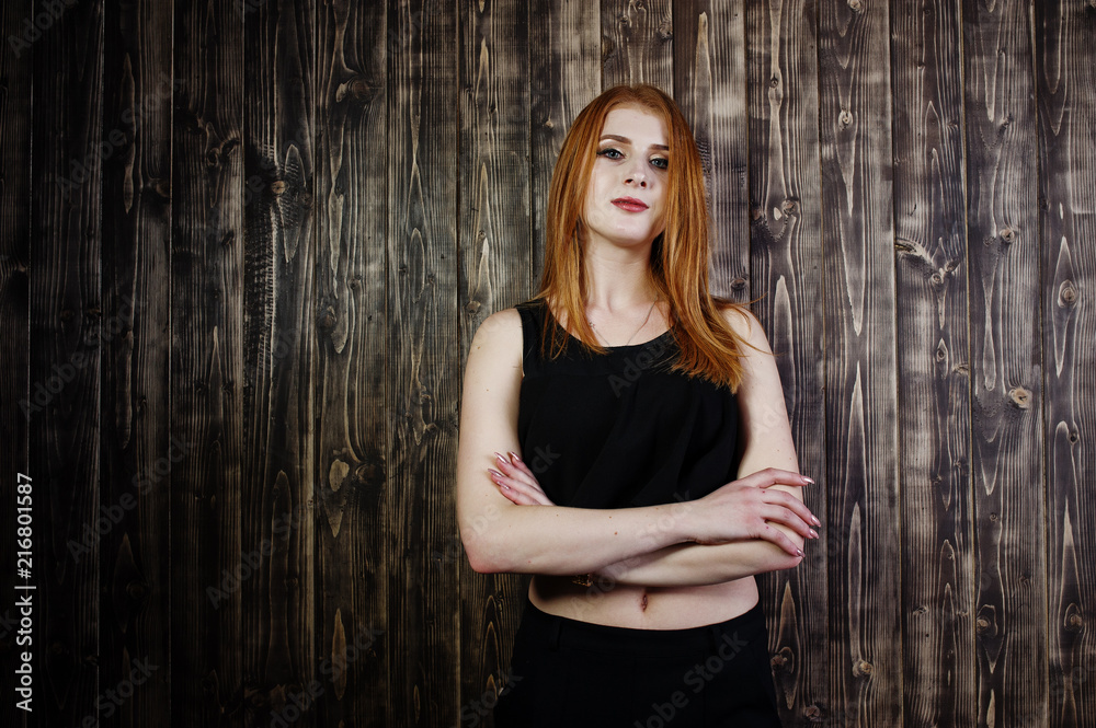 Fototapeta premium Portrait of a beautiful redheaded girl in black top and black skirt posing in the studio next to the wooden wall.
