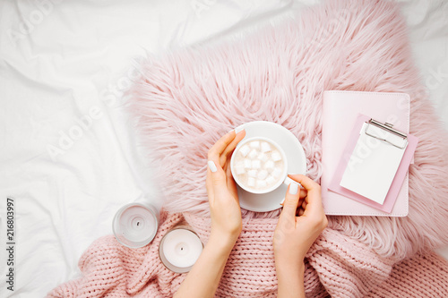 A cup of coffee on a pink pillow with notebook and a blanket on the bed. Flat lay, top view