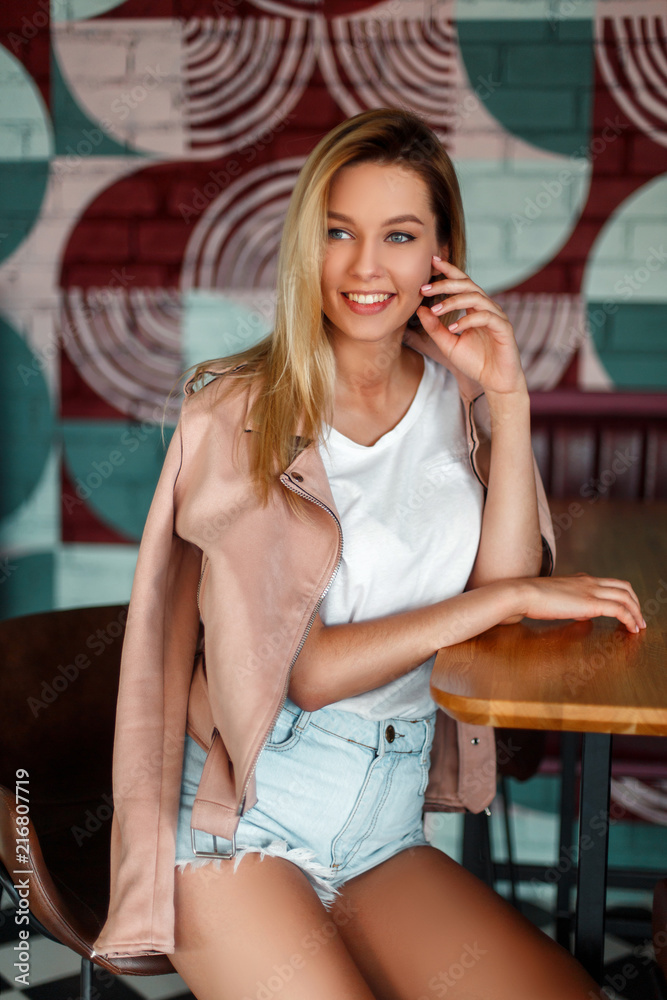 Beautiful happy woman model with a smile in a pink jacket in denim shorts  sitting on a chair at a table in a cafe Photos | Adobe Stock