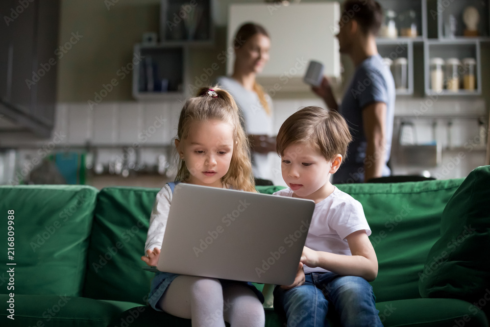 Cute children sister and brother watching online cartoons on laptop while  parents talking in the kitchen, curious siblings boy girl using pc at home,  kids computer addiction parental control concept Stock Photo |