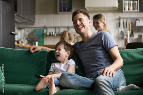 Happy dad and kid son holding remote control laughing at funny humor comedy film or tv show sitting on sofa at home, smiling father having fun with child boy watching television together on weekend