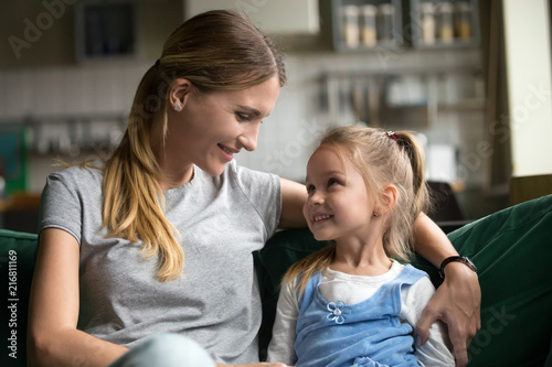 Smiling kid daughter looking at loving single mother hugging her on sofa, young woman mommy embracing cute happy girl showing care and support, sincere warm relationships of mom and child concept