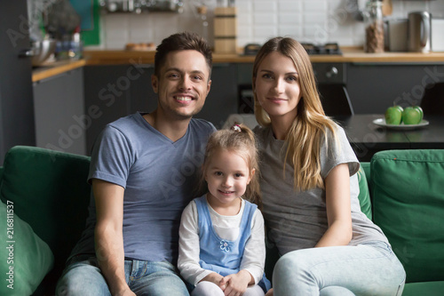 Portrait of happy healthy loving family of three concept, mom and dad posing with kid preschool daughter bonding together, young smiling parents sitting on sofa with child girl looking at camera