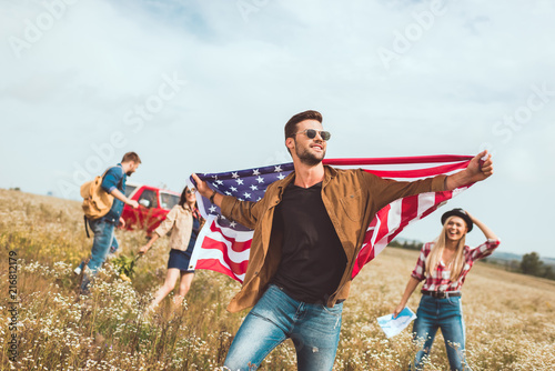 handsome young man holding usa flag and walking by field with friends during trip