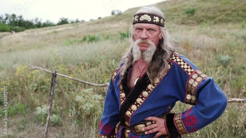 A view of an old tired man in ethnic costume and with gorgeous moustache stands holding hand on his waist. Slow motion photo